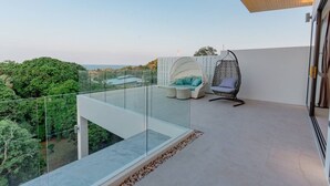 Un balcón con barandilla de vidrio, una silla colgante, una zona de estar con cojines y vistas a zonas verdes y edificios.