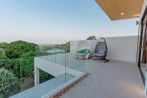 Ein Balkon mit Glasgeländer, einem Hängesessel, einer gepolsterten Sitzecke und Blick auf Grünflächen und Gebäude.