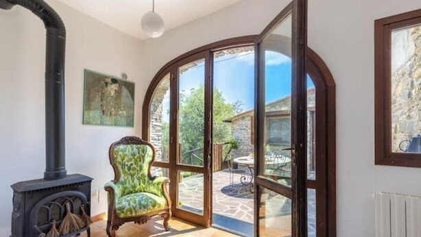 Une pièce dotée d’une cheminée, d’un fauteuil à motifs verts et d’un coin repas avec des sièges extérieurs, ainsi qu’une grande porte en verre en arc de cercle donnant sur un patio extérieur.