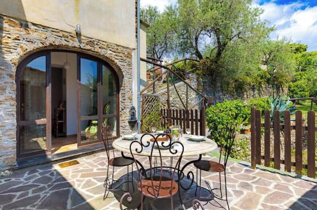 Un patio de piedra con una mesa redonda y sillas, un cerco de madera y un edificio con fachada de piedra y puertas de vidrio.