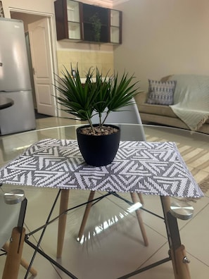 Ein Tisch mit Glasplatte, darauf ein Blumentopf, ein gemustertes Tischläufer und im Hintergrund eine Küchenzeile.