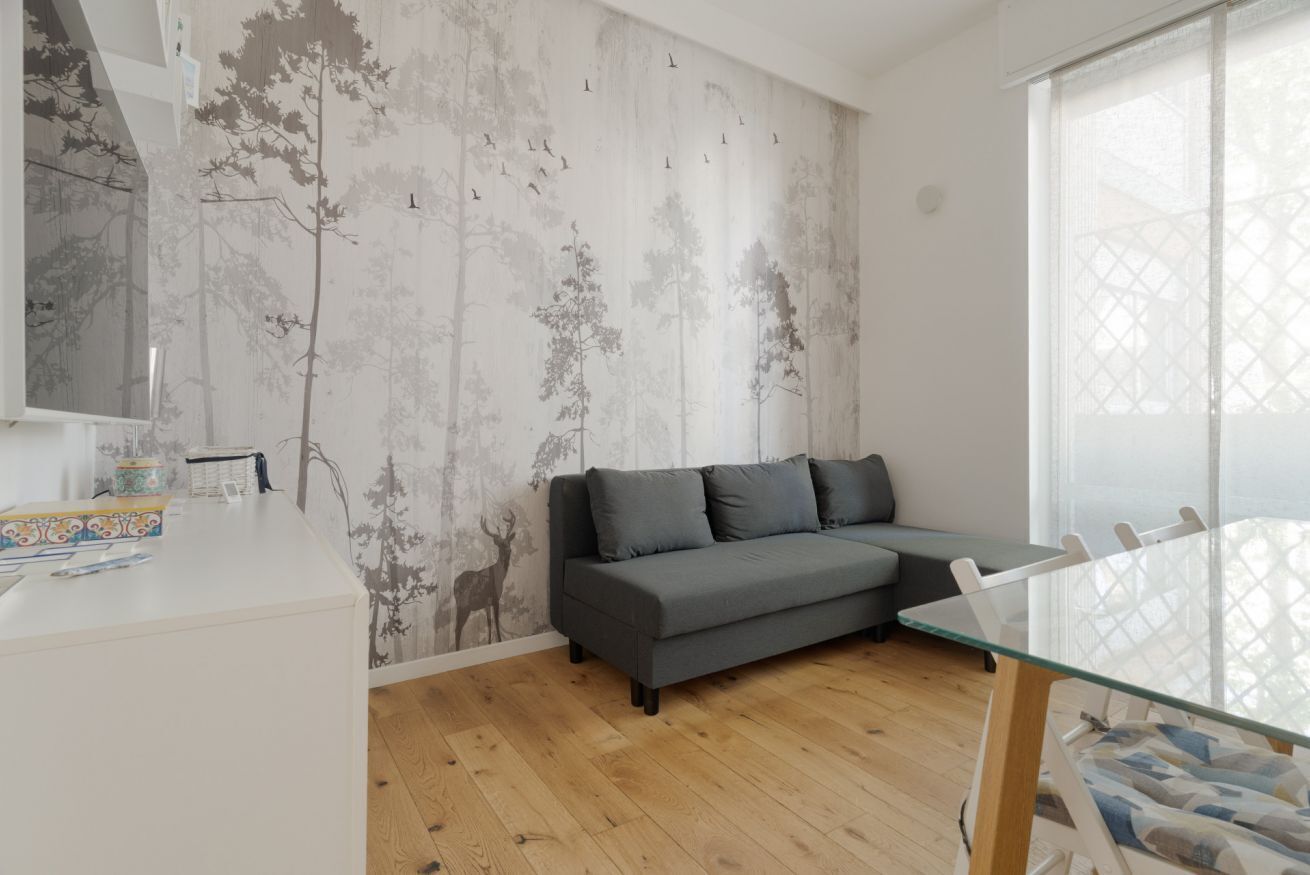 Un salón moderno con un sofá gris, una mesa de comedor de vidrio y una pared con un mural de un árbol y ciervos.