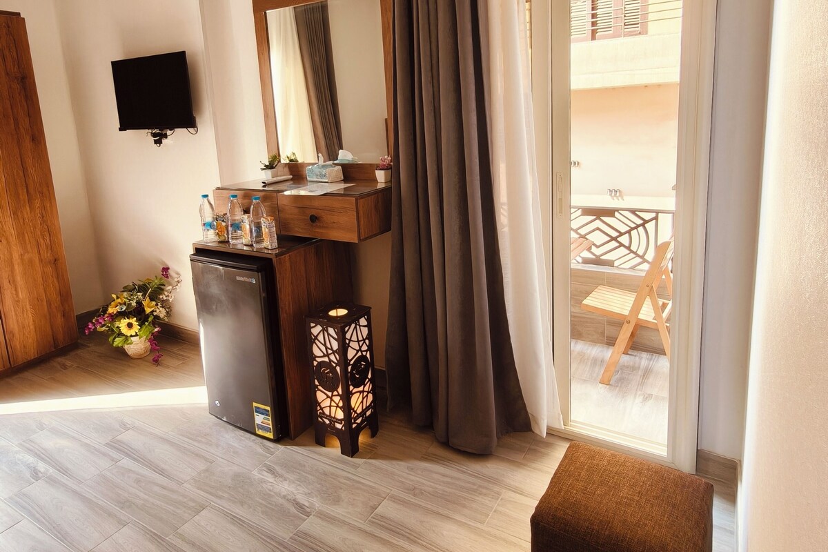 Una habitación con piso de madera, un escritorio con espejo, un refrigerador pequeño, un televisor montado en la pared y una puerta corrediza de vidrio que da a un espacio al aire libre.
