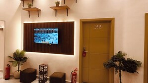 Um lobby moderno com televisão de tela plana, prateleiras de madeira com plantas, um extintor de incêndio e uma porta com a inscrição '103'.