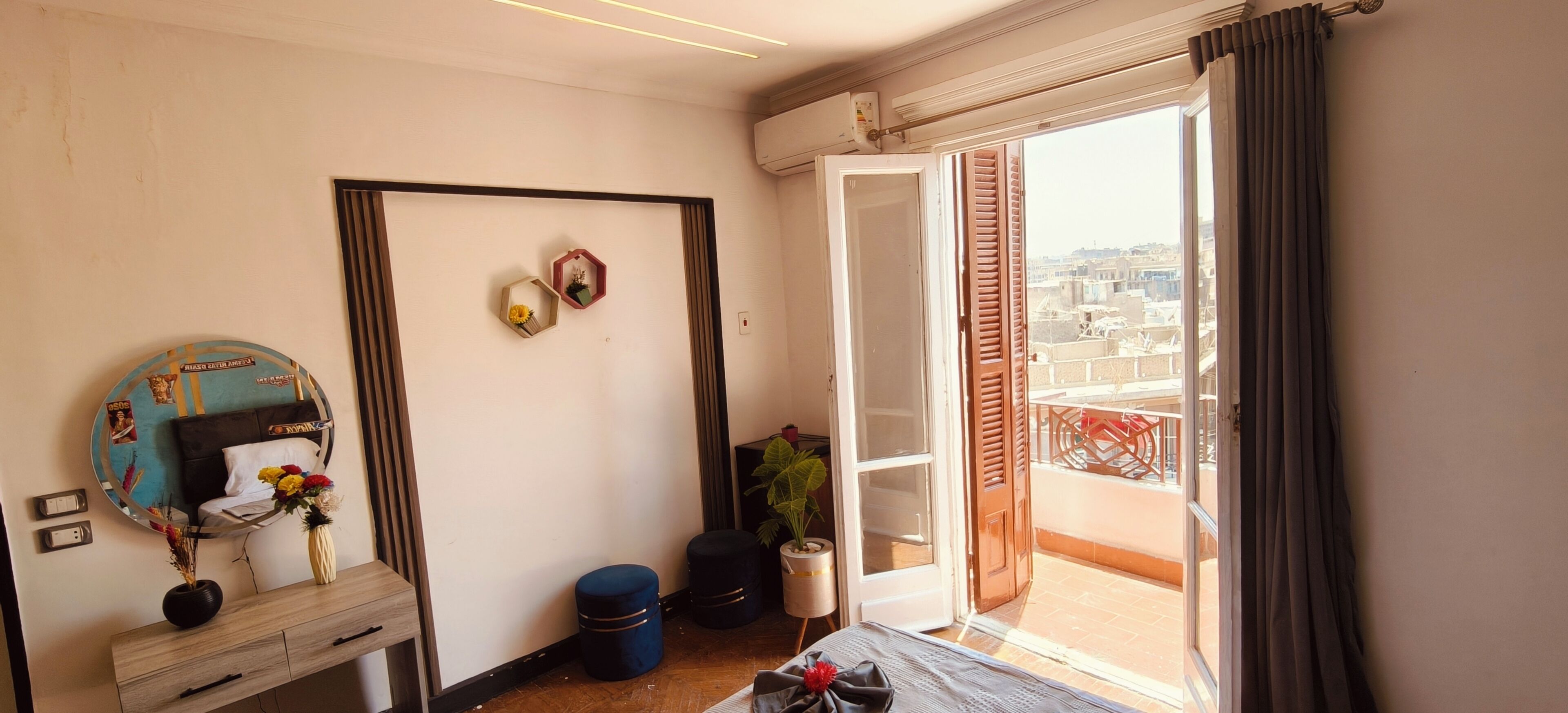 Quarto com cama, cômoda, espelho, janela com vista para edifícios e porta que dá para uma varanda.