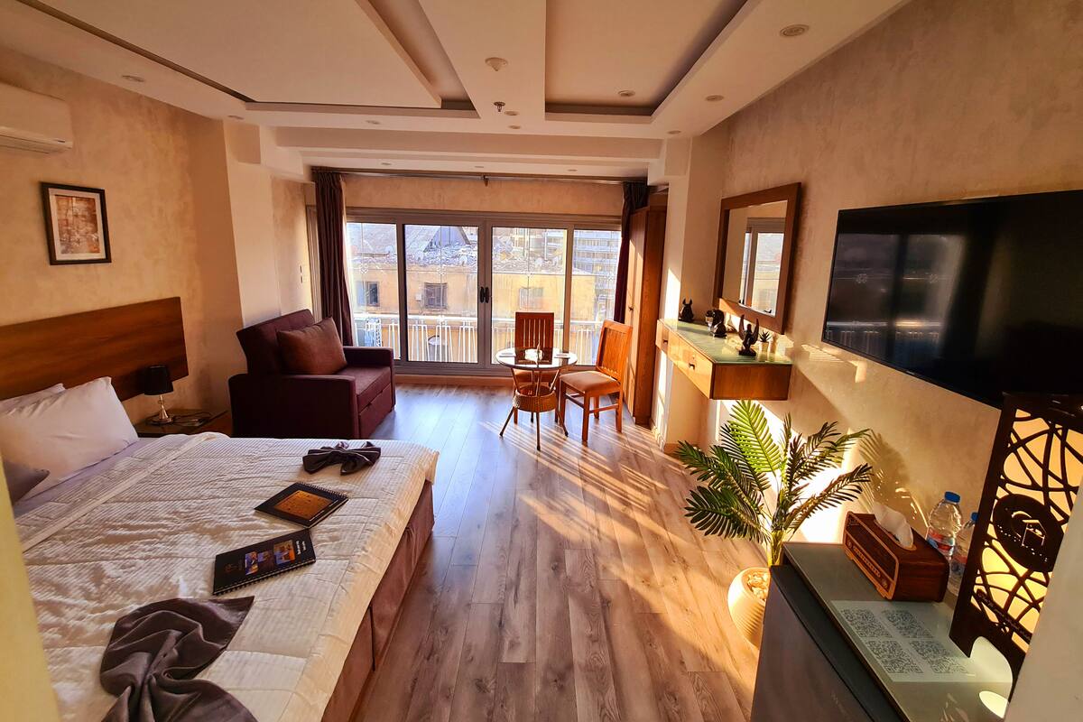 A hotel room with a bed, a sofa, a dining table with chairs, a TV, and a balcony with a view.