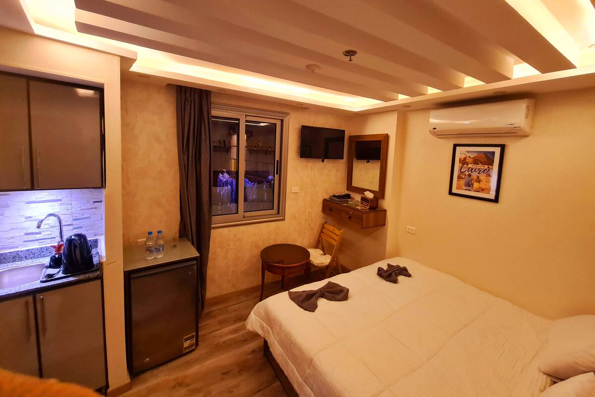 A compact hotel room with a kitchenette, a bed, a small desk, a chair, a window, and a mounted TV.
