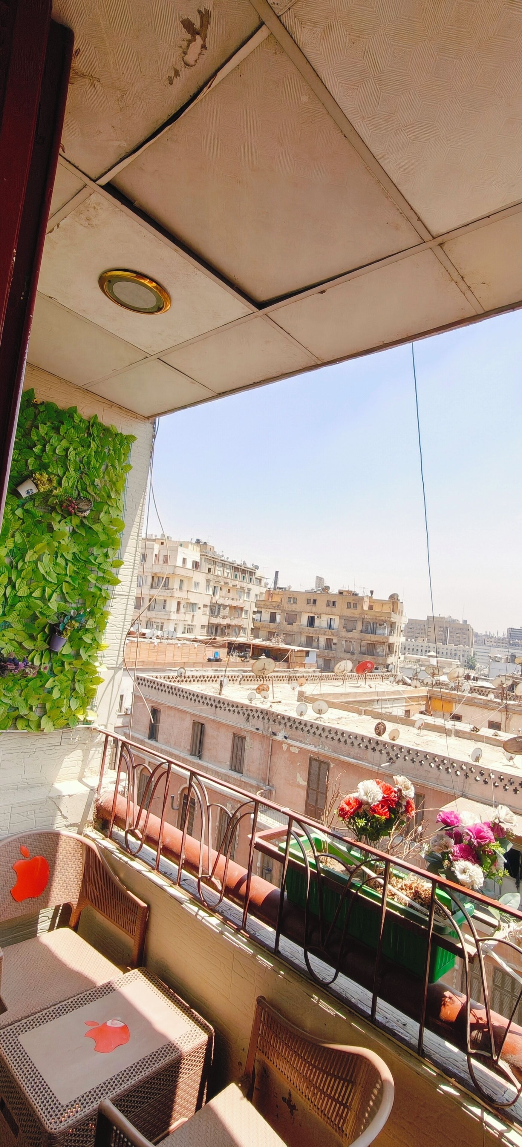 Un balcón con vistas a un paisaje urbano, que incluye barandilla, plantas en macetas y una mesa con una silla.