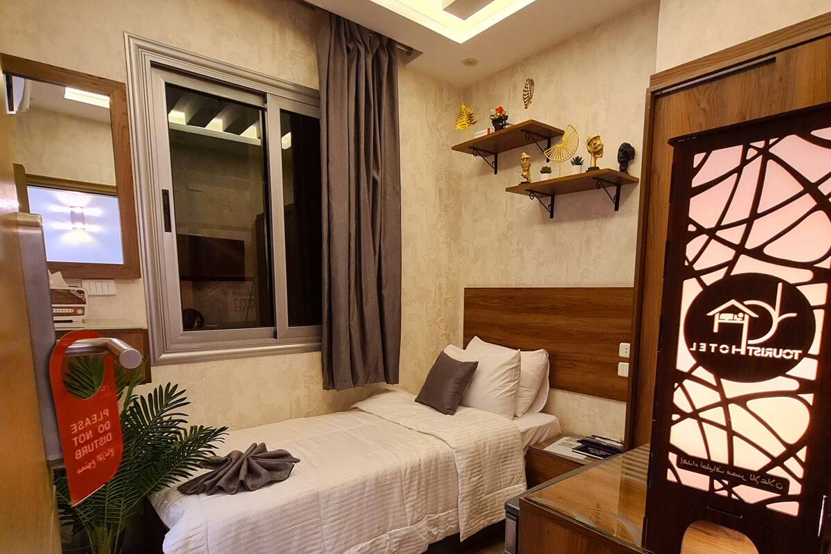 A hotel room with a bed, a desk, a potted plant, a window with curtains, a shelf with decorative items, and a wall-mounted TV.