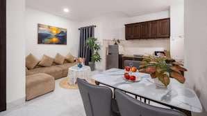 A modern living room with a sofa, a dining table with chairs, a kitchen area with cabinets, and a painting on the wall. - Miami Beach Hotel & Apartment (Da Nang)
