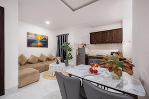 A modern living room with a sofa, a dining table with chairs, a kitchen area with cabinets, and a painting on the wall. - Miami Beach Hotel & Apartment (Ngu Hanh Son)