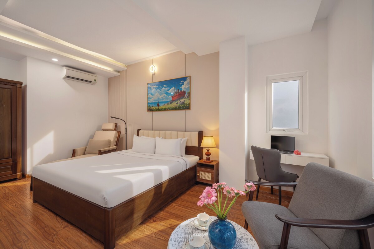 A hotel room with a large bed, a desk with a computer, a chair, and a small table with a vase of flowers.