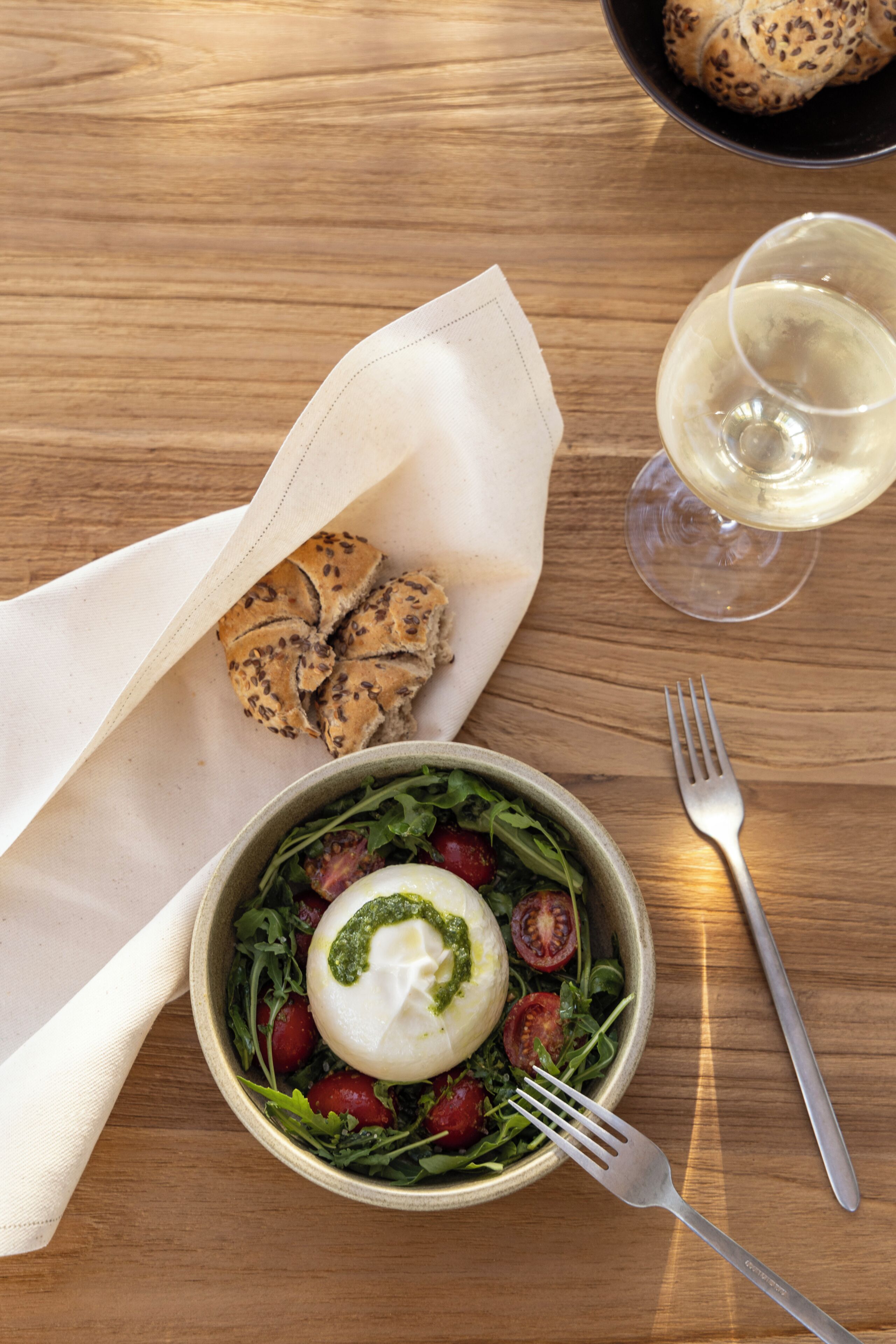 A wooden table with a bowl of salad, a glass of white wine, a fork, and a napkin.