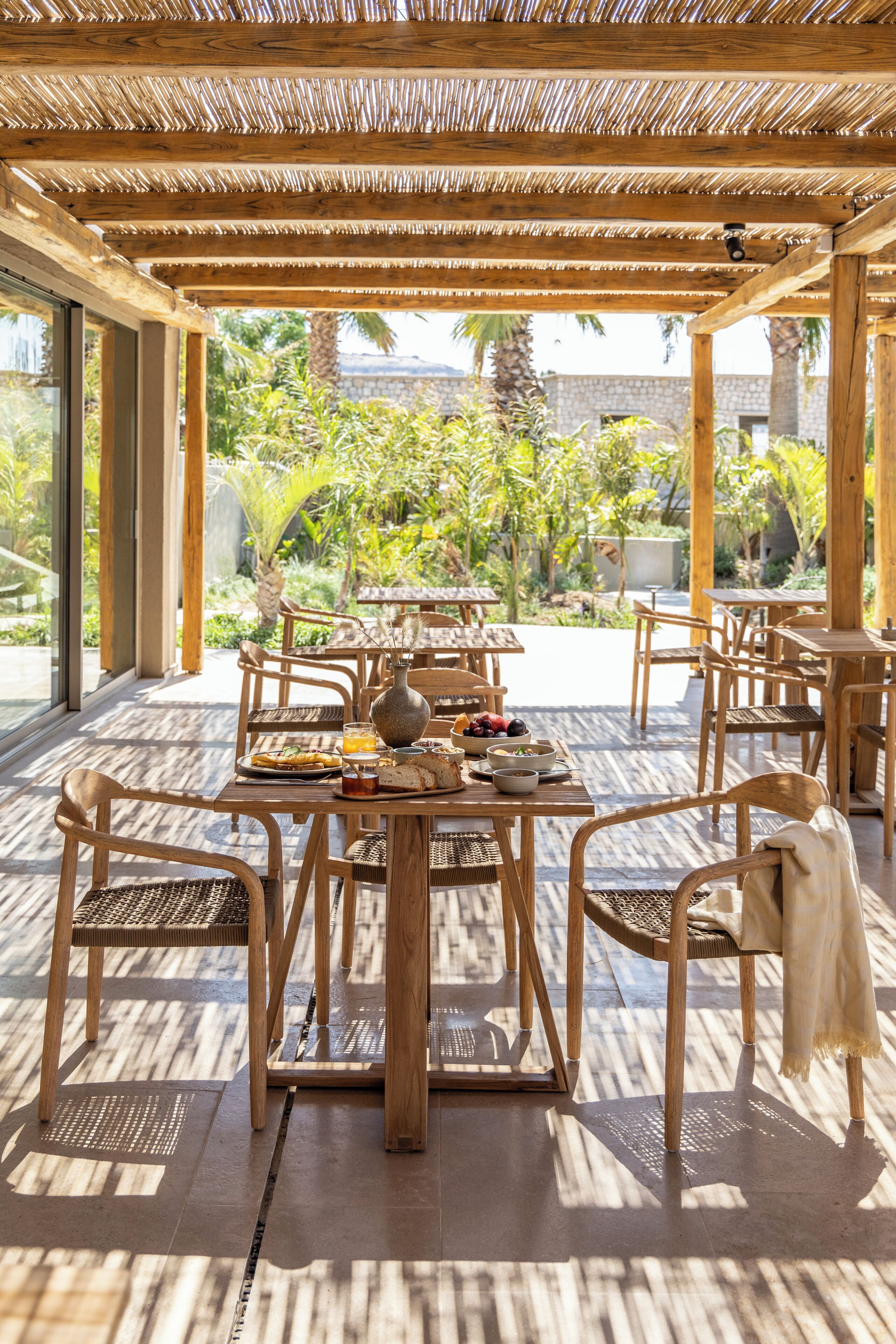 Ein Essbereich im Freien mit Holztischen und -stühlen, umgeben von einer Pergola mit einem Reetdach.