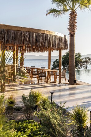 A wooden outdoor seating area with tables and chairs, a thatched roof structure, and a palm tree. - Pefkian Ethos Hotel & Suites (Rhodes)