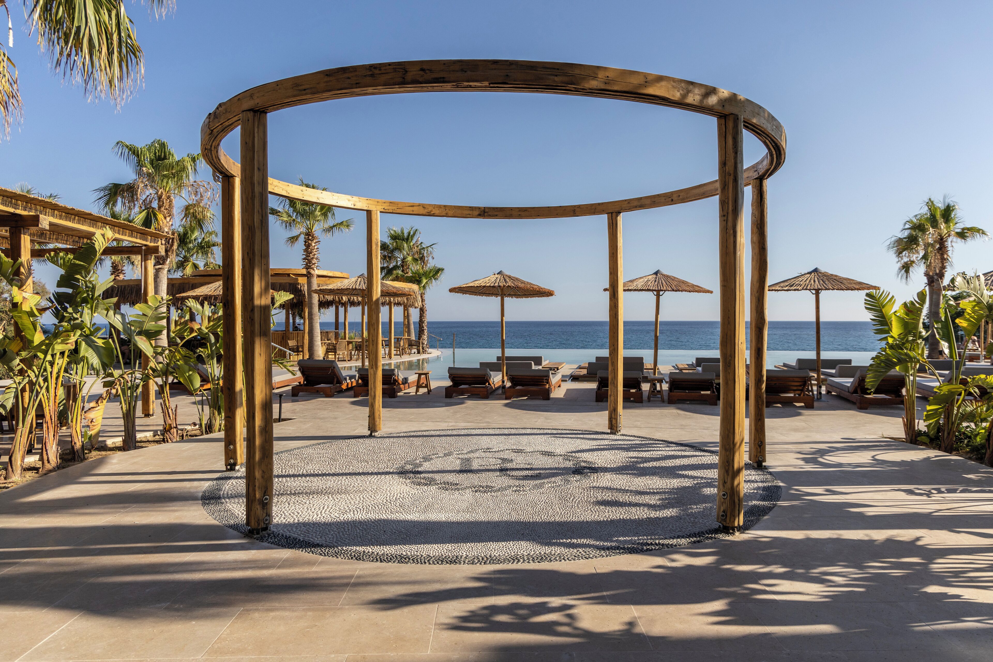 Una estructura de madera tipo quiosco con asientos, palmeras y vista a la playa.