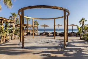 Una estructura de madera tipo quiosco con asientos, palmeras y vista a la playa.