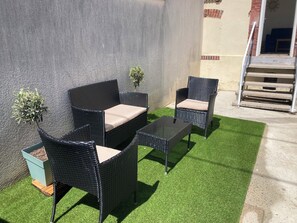 Eine Terrasse mit Korbmöbeln, einem Glastisch und kleinen Kübelpflanzen.