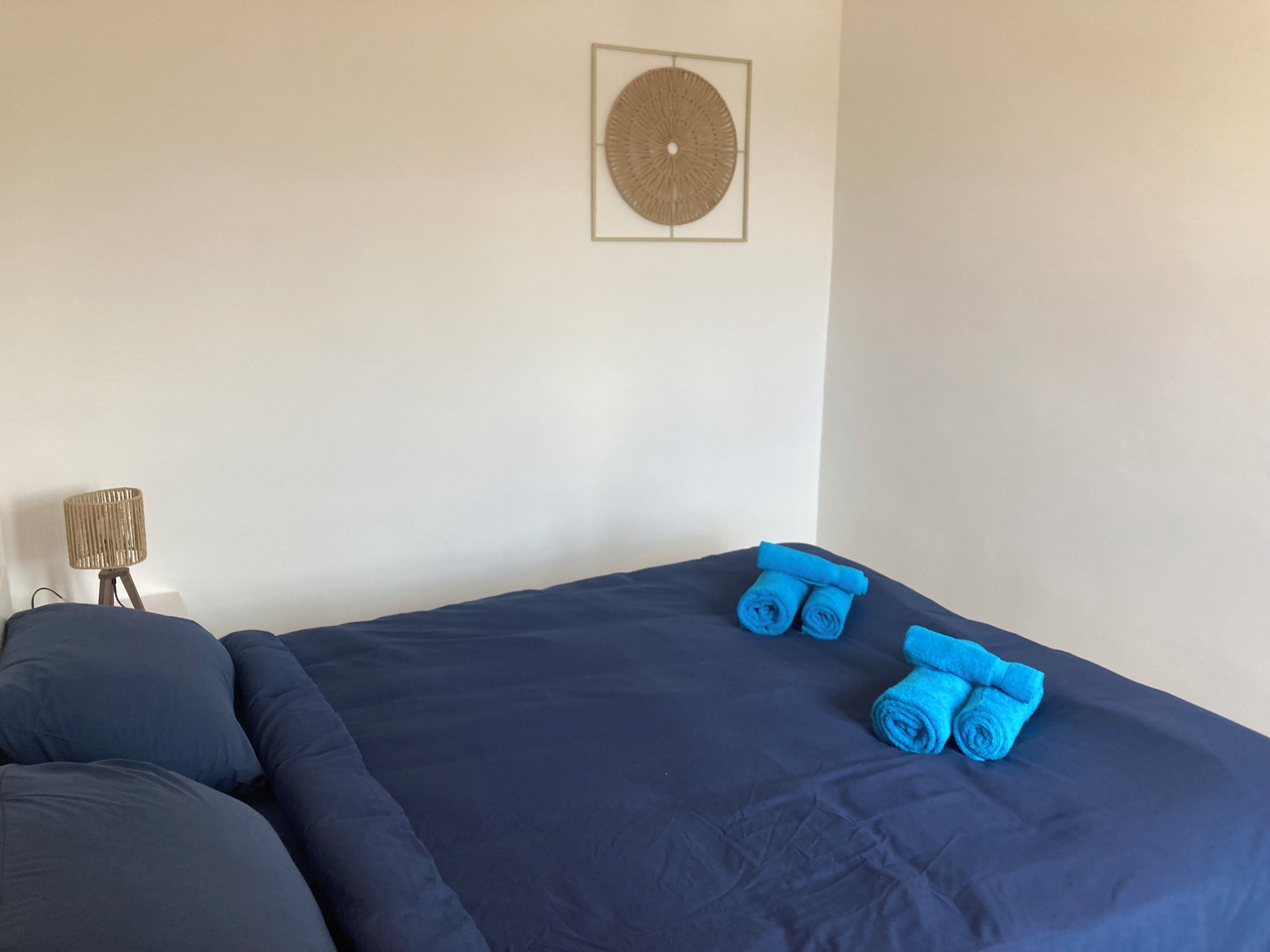 A bed with dark blue bedding, two rolled towels, a lamp, and a wall-mounted decorative item.