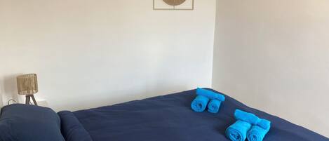 A bed with dark blue bedding, two rolled towels, a lamp, and a wall-mounted decorative item.