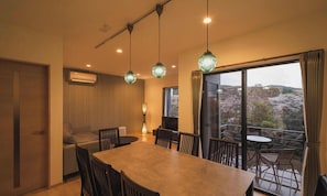 Un comedor con una mesa grande, sillas, lámparas colgantes verdes, un sofá y un televisor.