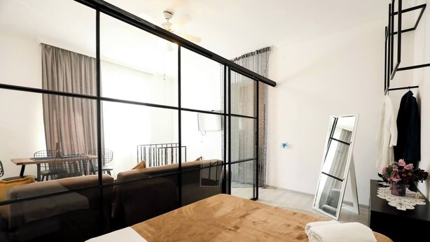 A hotel room with a large bed, a desk with a chair, a mirror, and a view through a glass partition.