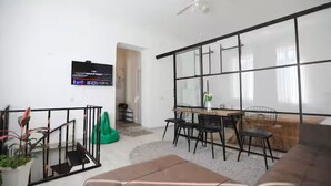 A modern living room with a sofa, a dining table with chairs, a TV mounted on the wall, and a large window with a view.