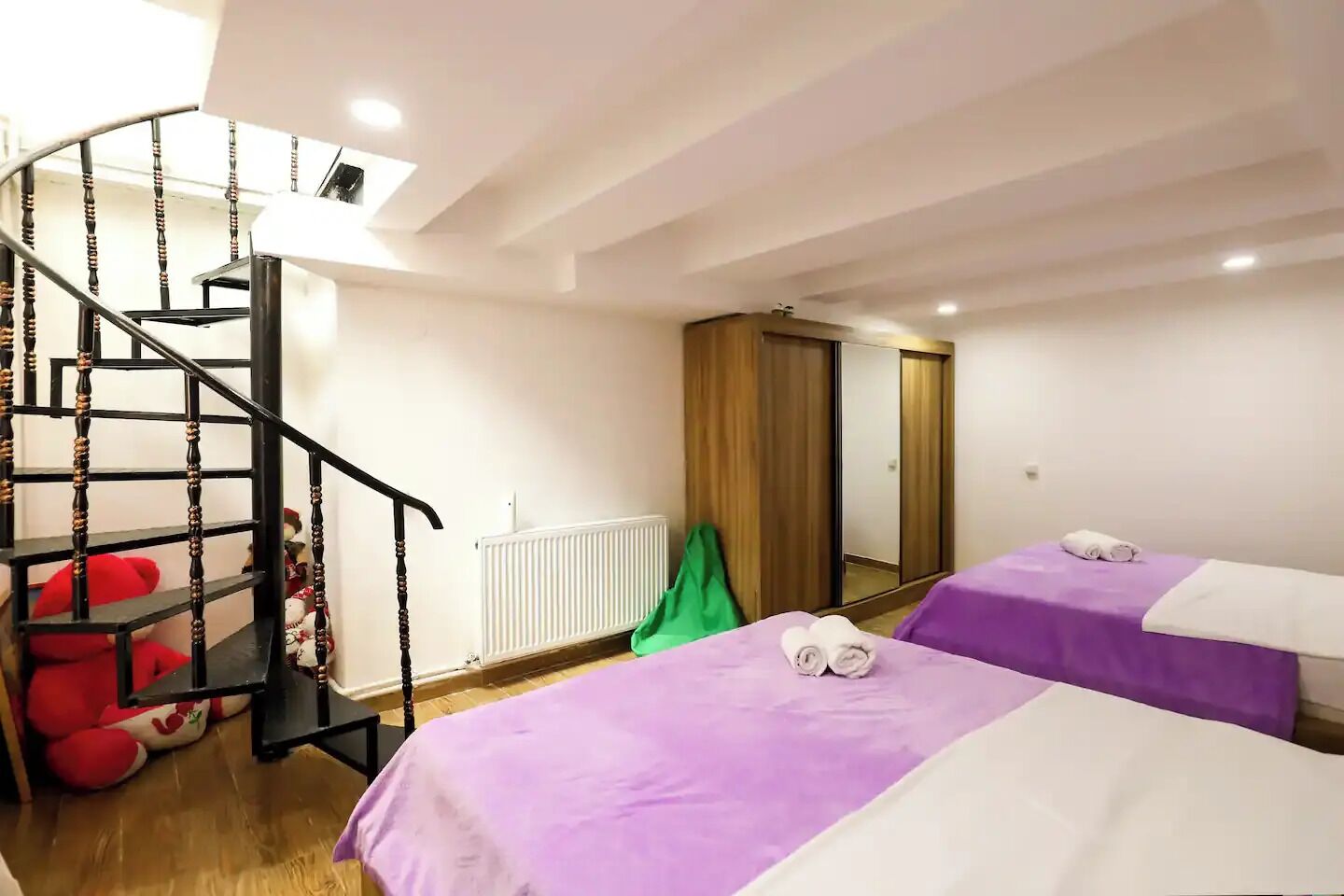A hotel room with a spiral staircase, two beds with purple and white bedding, a radiator, and a mirror on the wall.