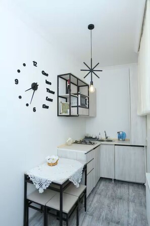 Une cuisine compacte avec une horloge murale blanche, un luminaire suspendu moderne et une petite table à manger recouverte d’une nappe en dentelle blanche.