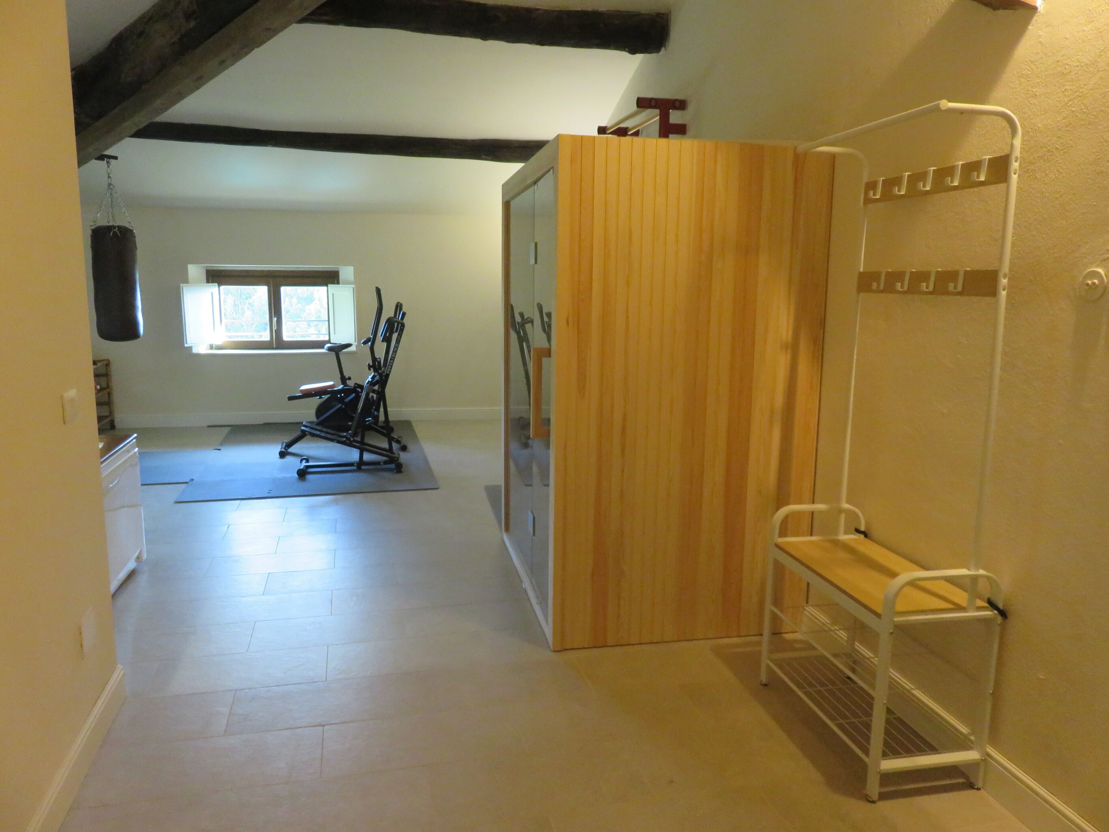 A room with a wooden wardrobe, a white metal bench, an exercise machine, and a window with natural light.