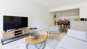 A modern living room with a TV, a round coffee table, a sofa, and a dining area with chairs and a kitchen in the background. - Quartier Carlton 3 rooms South with terrace (Cannes)