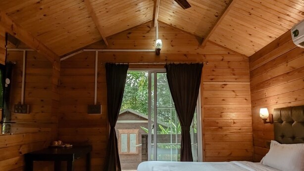 A wooden cabin bedroom with a single bed, a ceiling fan, a small table, and a window with curtains. - Greenland cottage (Arambol)