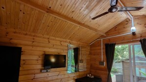 A wooden cabin interior with a ceiling fan, a mounted TV, a small table with a tray, and a sliding glass door leading outside. - Greenland cottage (Arambol)