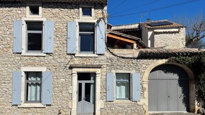 Ein Steinhaus mit zahlreichen Fenstern und Fensterläden, einem großen Bogenportal und einem kleinen Anbau mit Ziegeldach.
