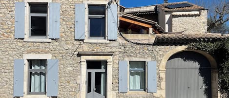 Un edificio de piedra con múltiples ventanas y contraventanas, una gran entrada en arco y una estructura anexa pequeña con tejado de tejas.
