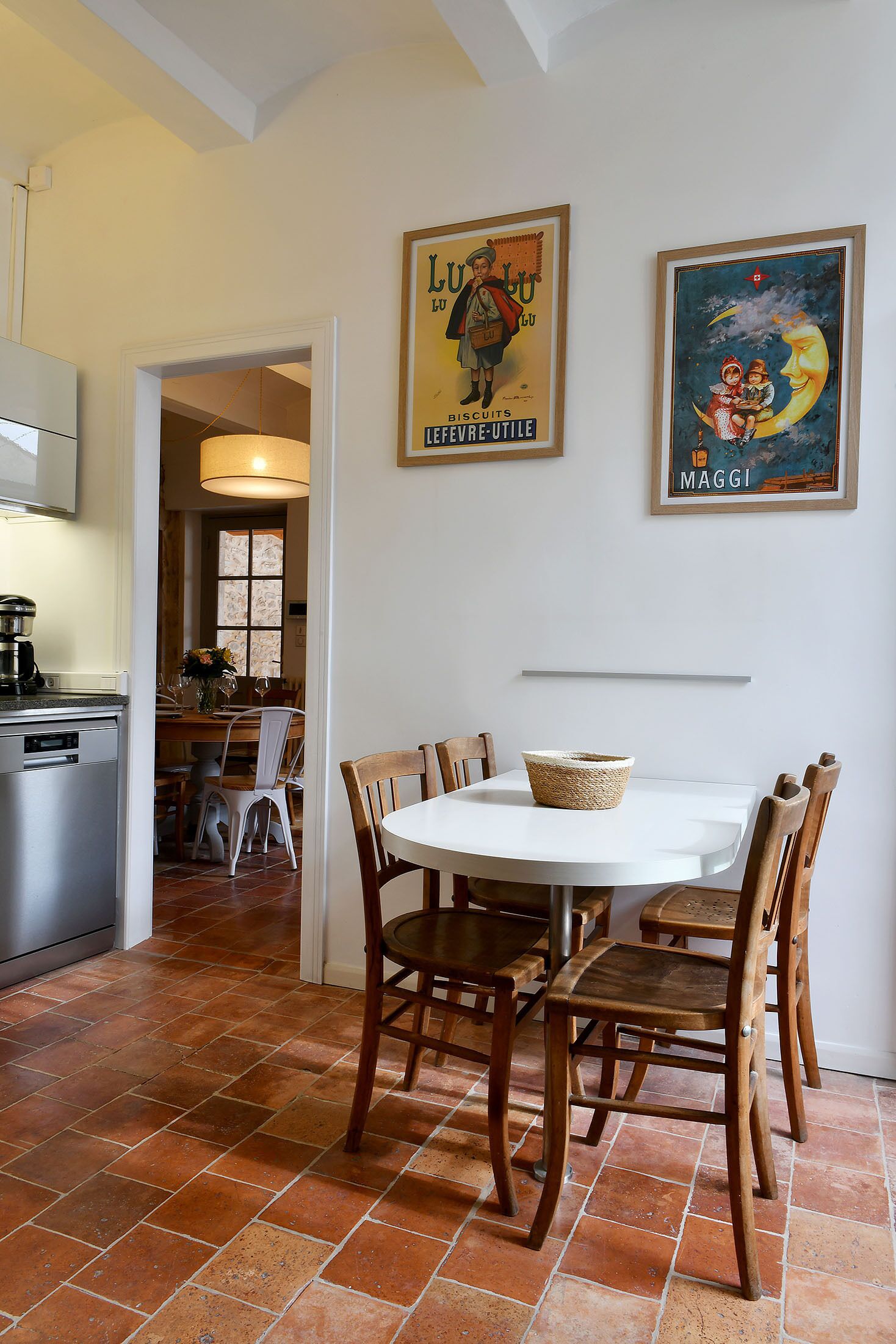 A dining area with a round table, wooden chairs, and a white tablecloth. There are two framed pictures on the wall, a kitchen with a stove, and a window with curtains.