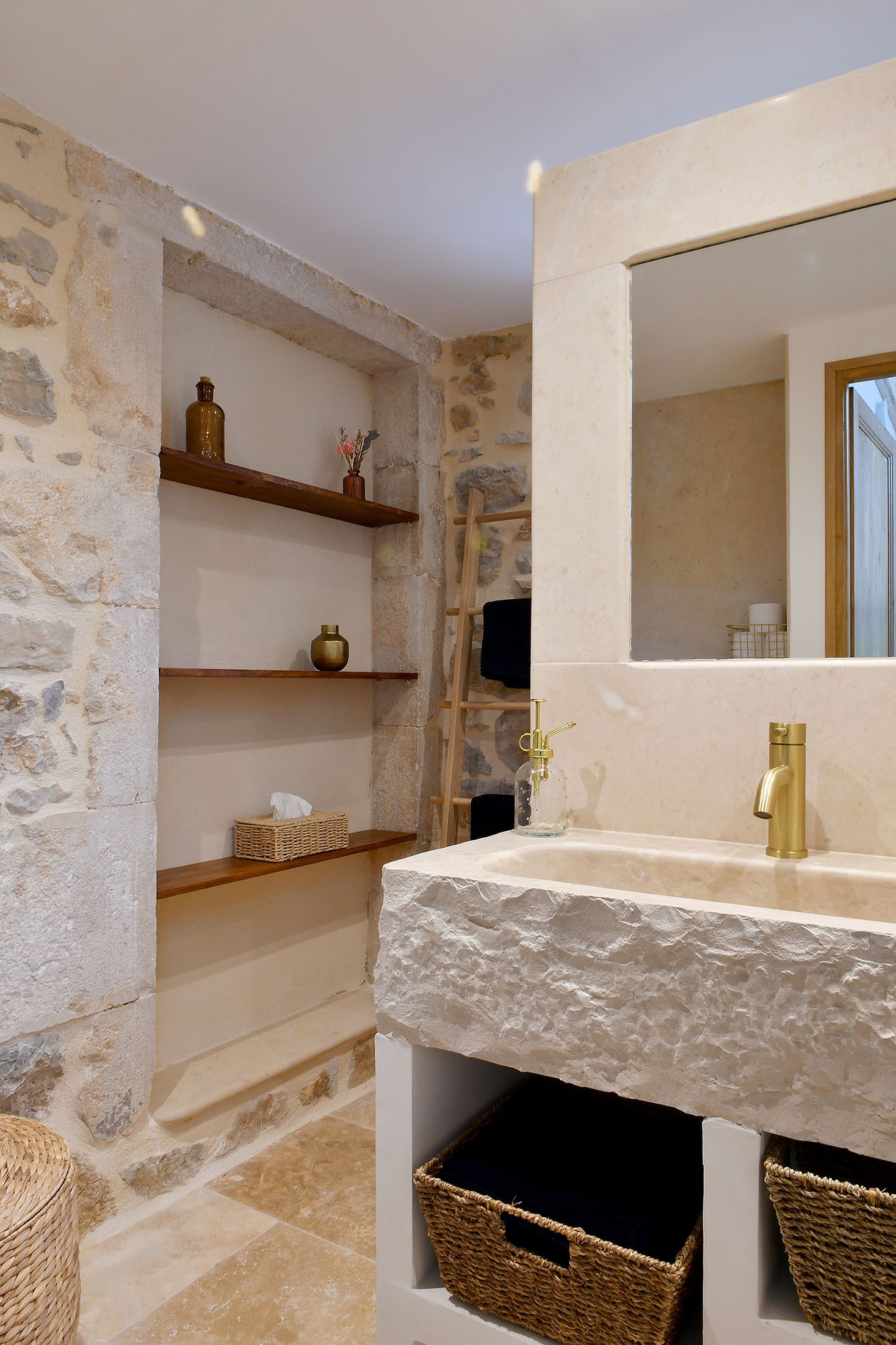 A bathroom with stone walls, a wooden shelf with decorative items, a gold faucet, and wicker baskets under the sink.