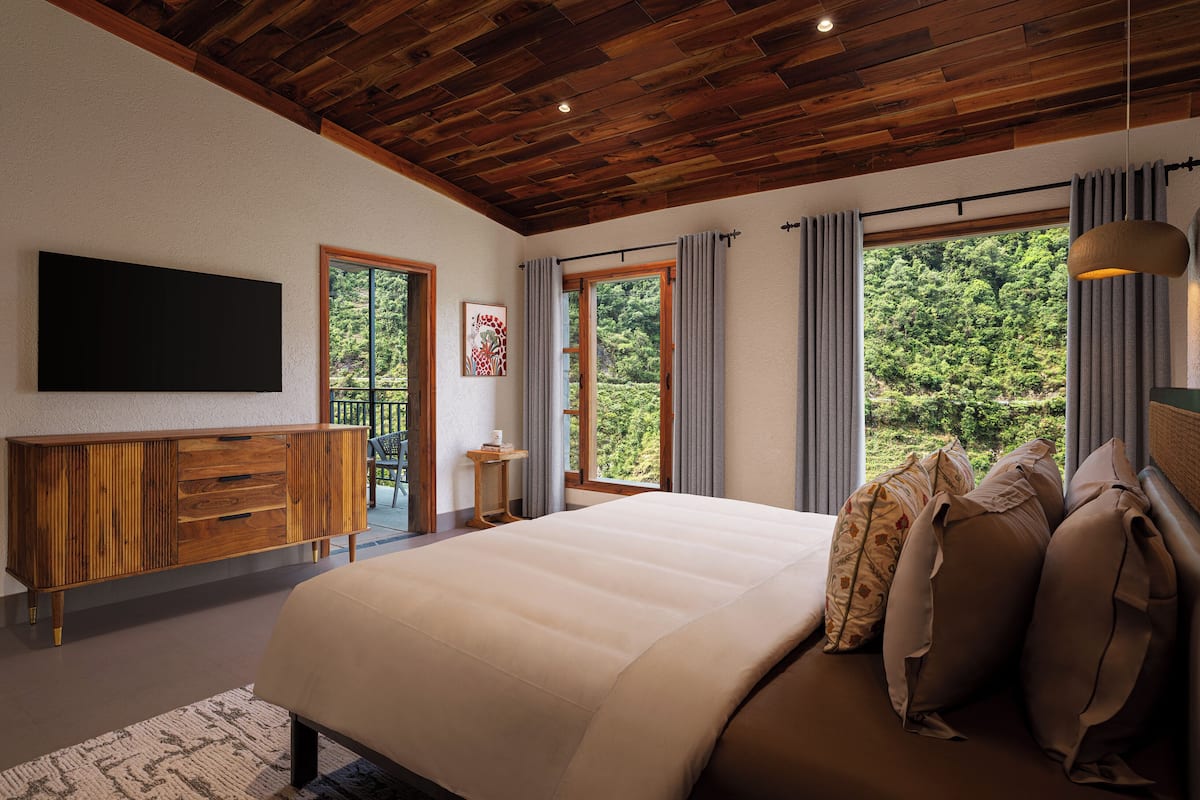 Une chambre avec un plafond en bois, un grand lit, une télévision et une vue sur la verdure par les fenêtres.