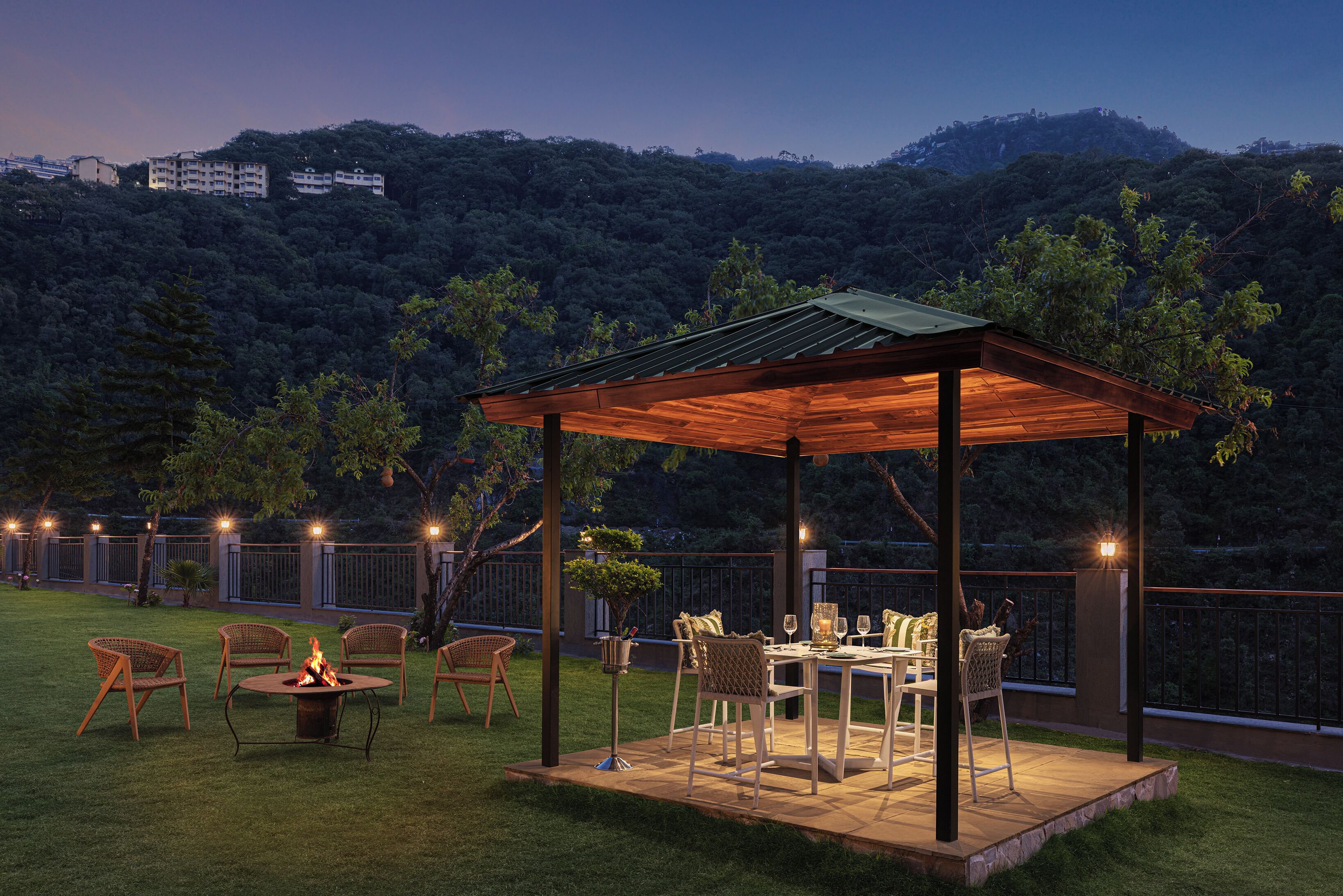 A covered outdoor seating area with a fire pit, chairs, and a table set for dining, surrounded by a lush garden and a view of a hill with buildings.