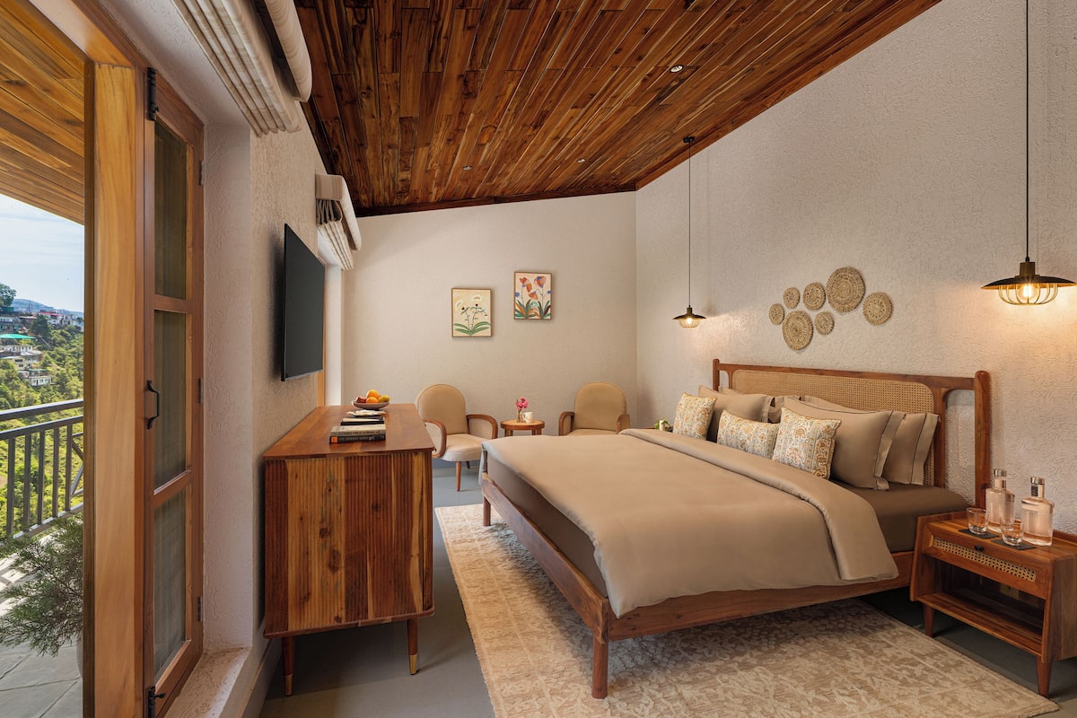 Une chambre avec un plafond en bois, un lit avec une literie beige, un bureau, deux chaises, une télévision et un balcon avec vue.