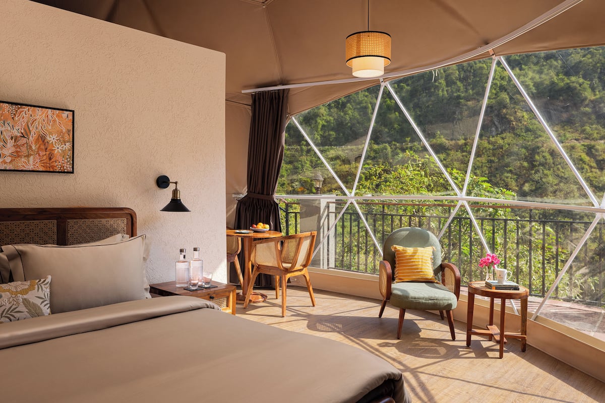 Une chambre à coucher comprenant un lit, une table de chevet, une chaise, une petite table avec un vase, et une vue dégagée sur l’extérieur grâce à une grande fenêtre.