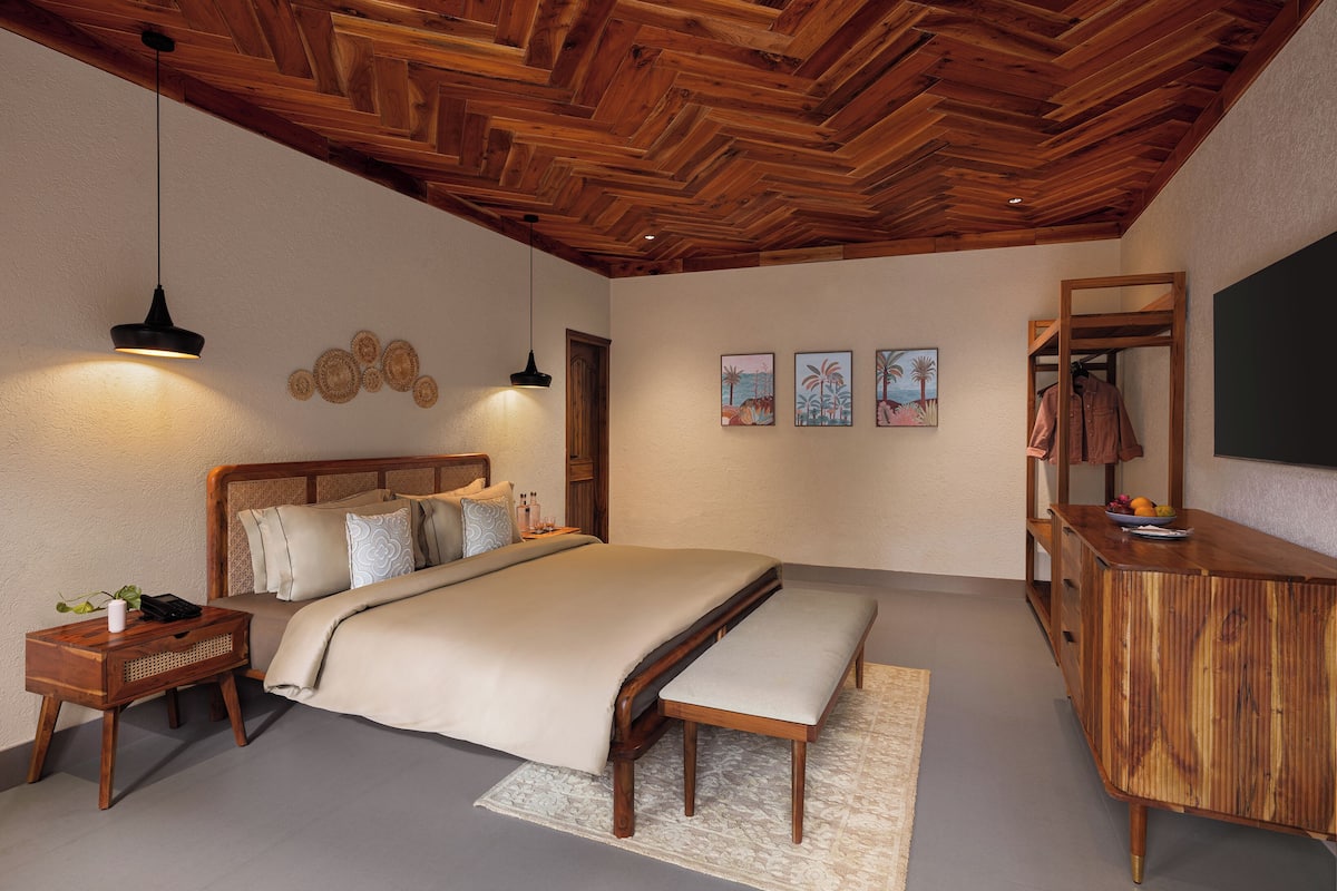 Une chambre avec un plafond en bois, un lit avec une banquette, des tables de chevet, des œuvres d’art murales et une commode en bois.