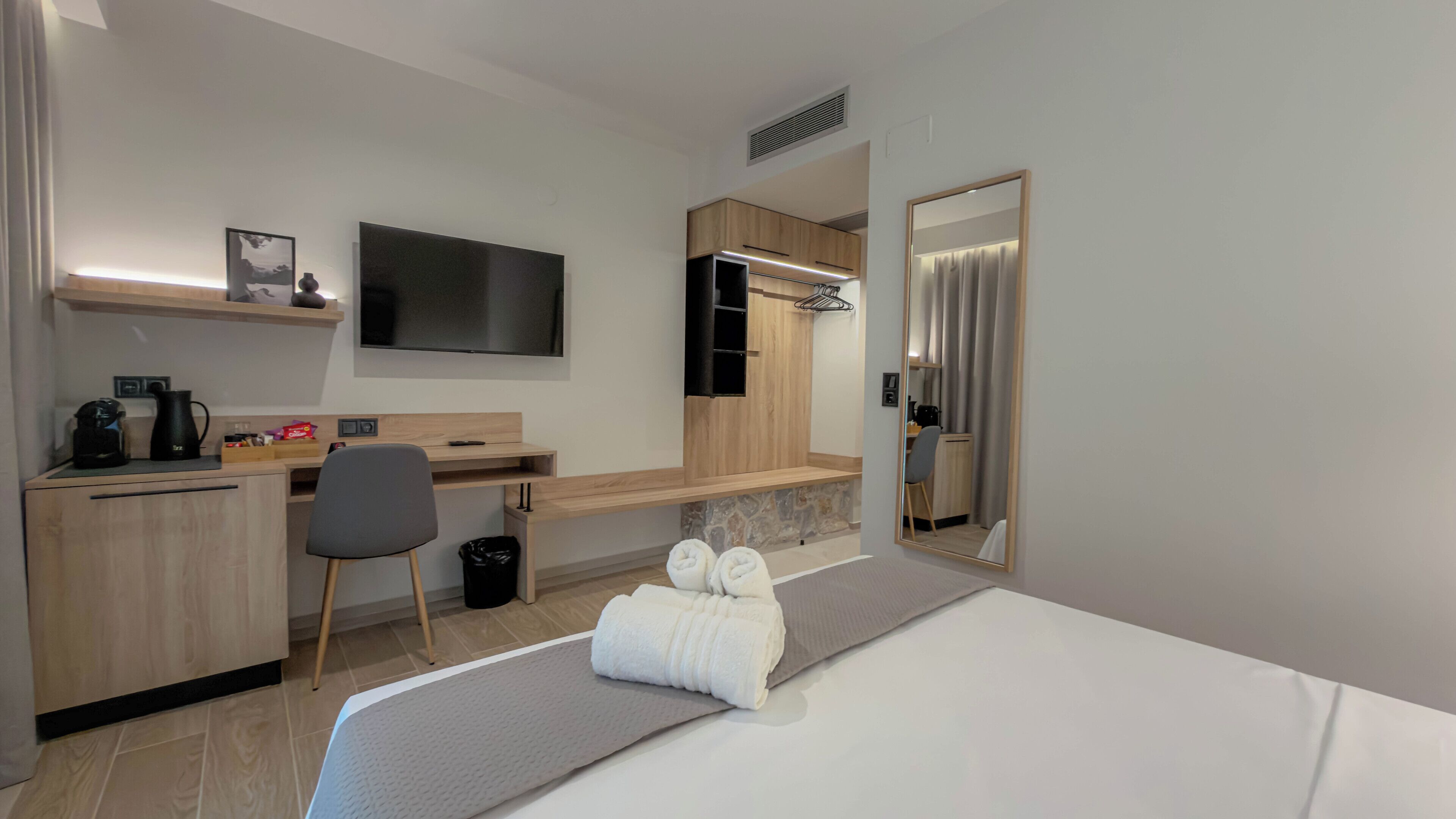 A hotel room with a bed, a desk, a chair, a TV, a mirror, and a small shelf with a picture.