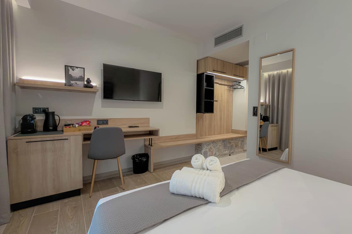 A hotel room with a bed, a desk, a chair, a TV, a mirror, and a small shelf with a picture.