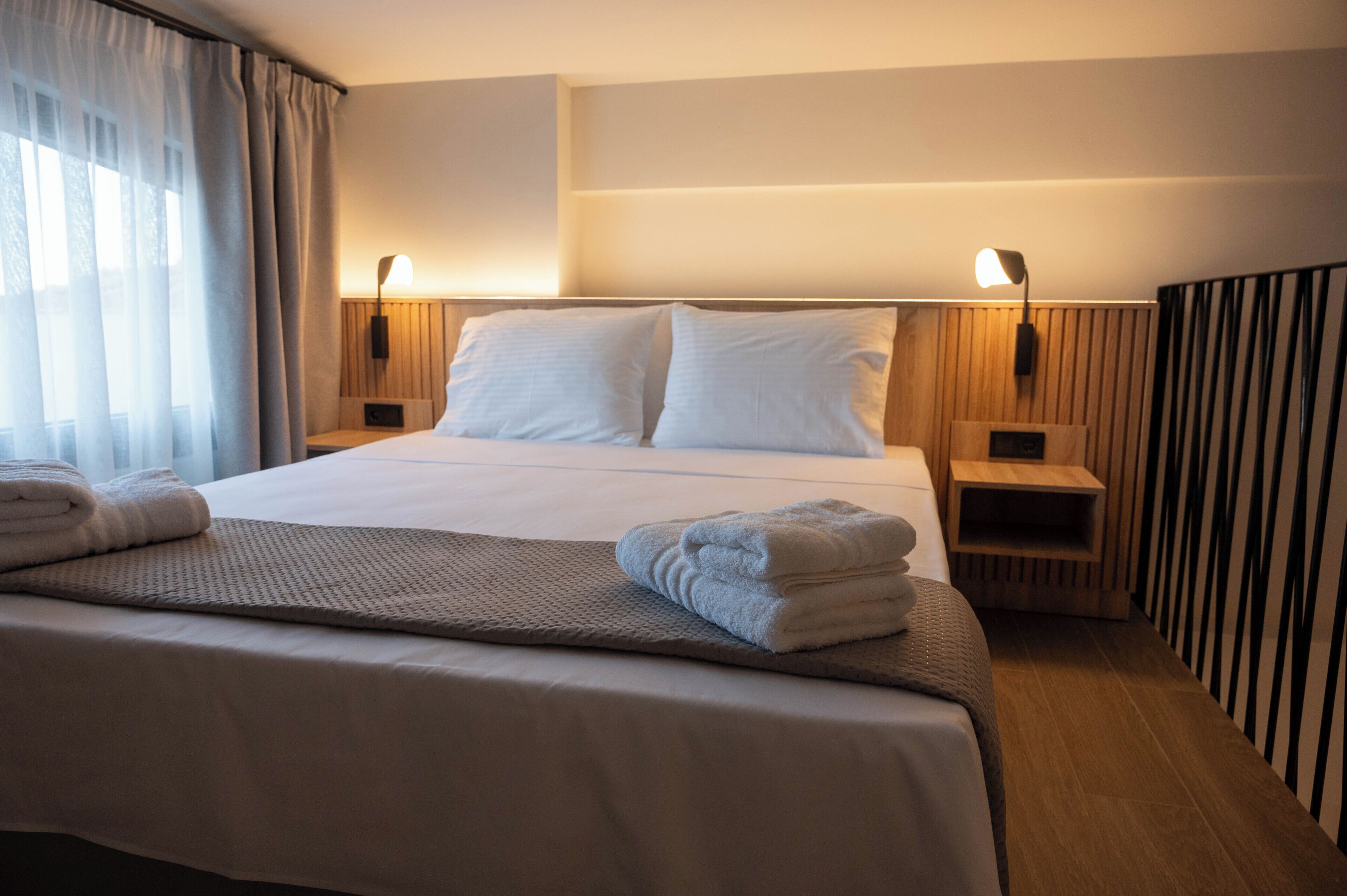 Una habitación de hotel con una cama bien hecha, dos mesitas de noche con lámparas, un cabecero de madera y un gran ventanal con cortinas.