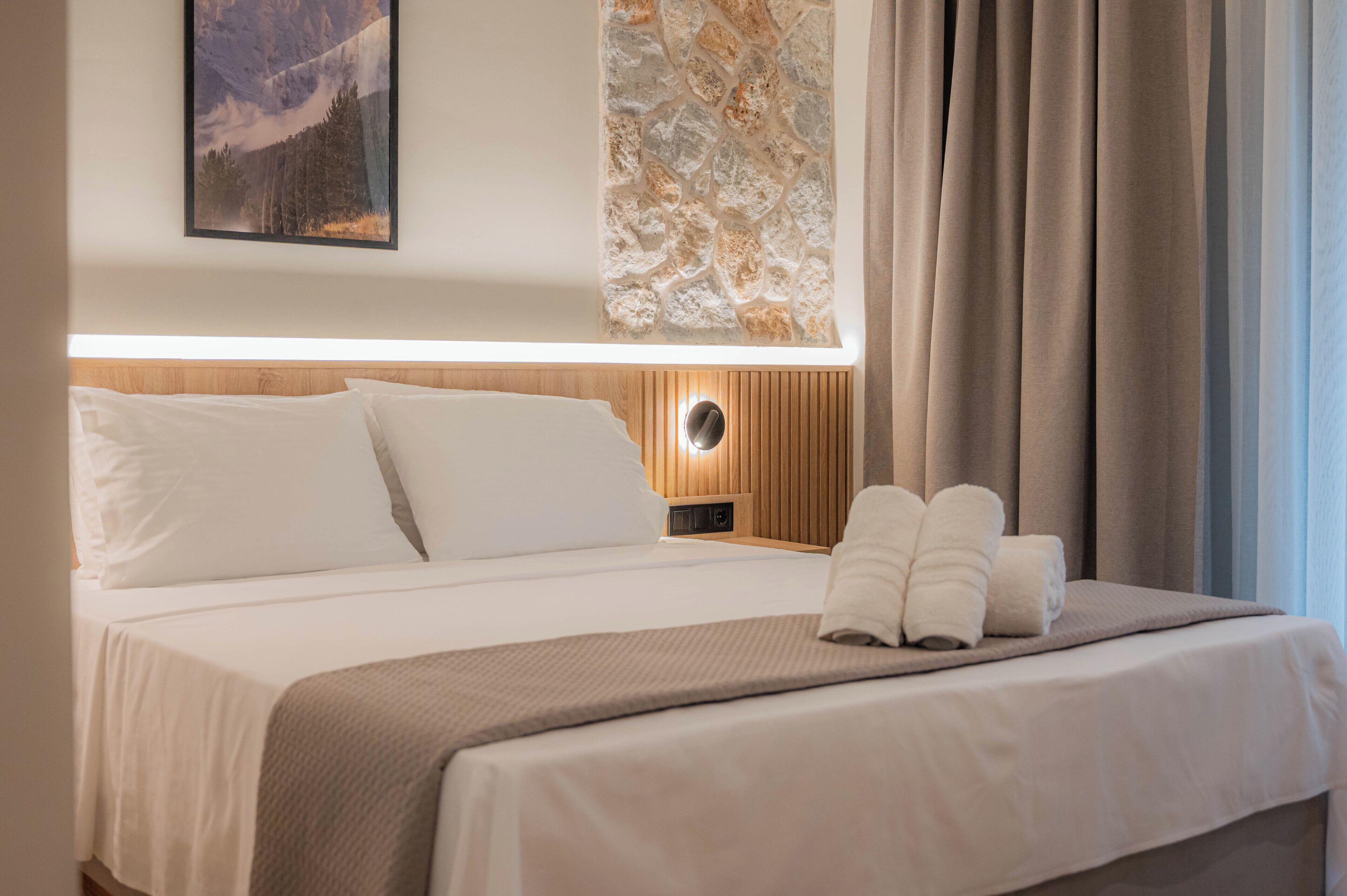 Un lit bien fait, avec du linge de lit blanc, une tête de lit en bois, une lampe fixée au mur et un tableau encadré au-dessus.