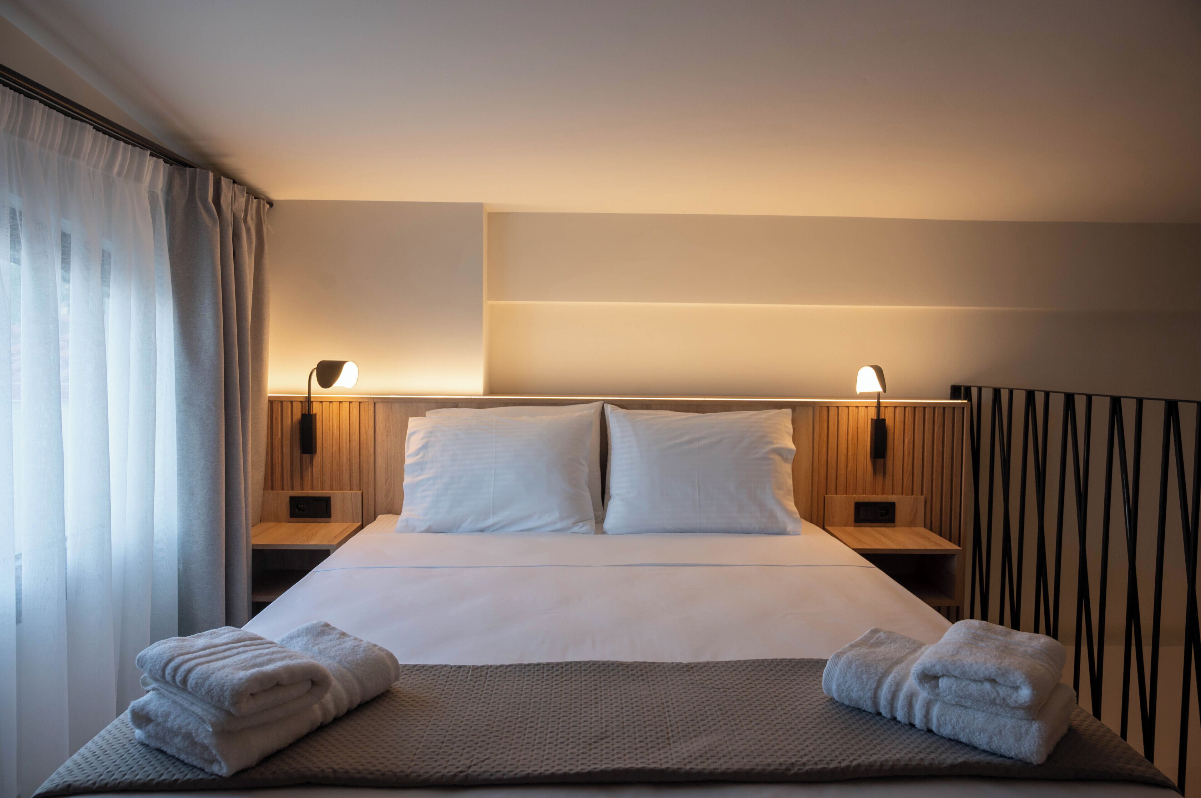 A hotel room with a neatly made bed, two bedside lamps, a wooden headboard, and a window with curtains.