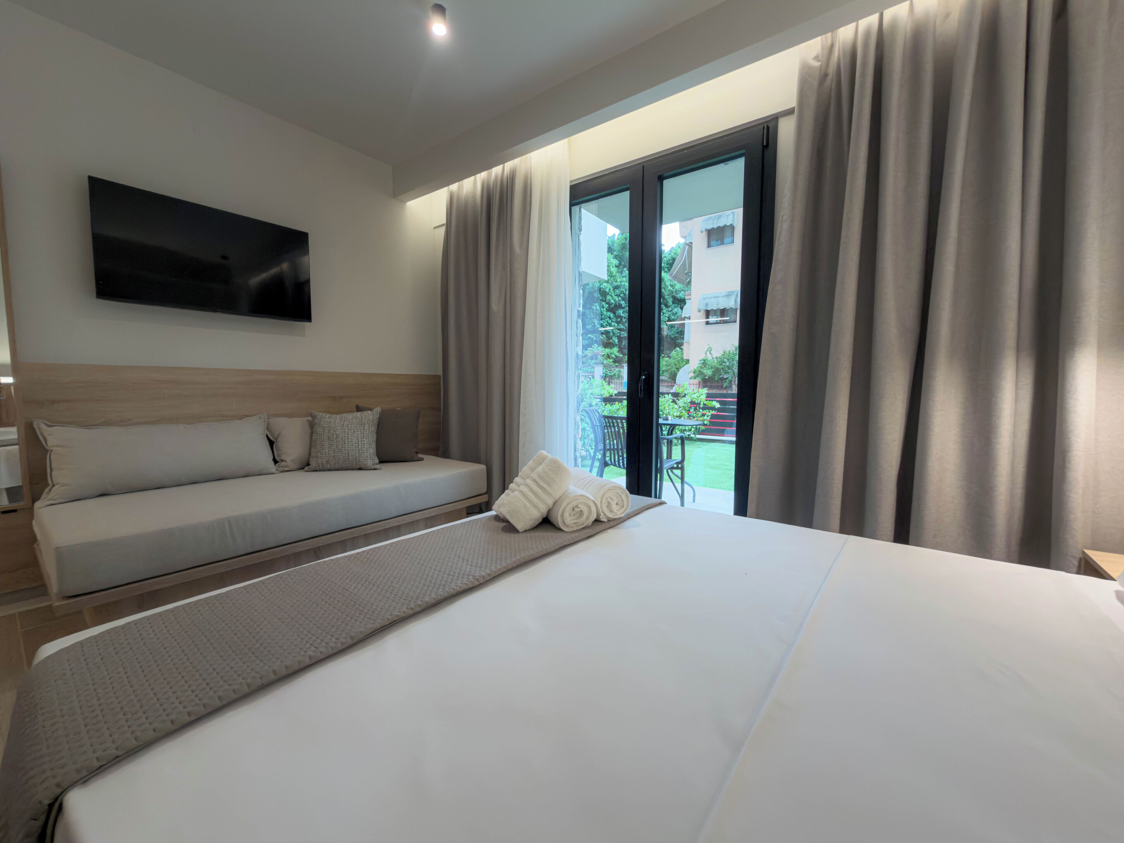 A hotel room with a bed, a sofa, a TV mounted on the wall, and a view of the outside.