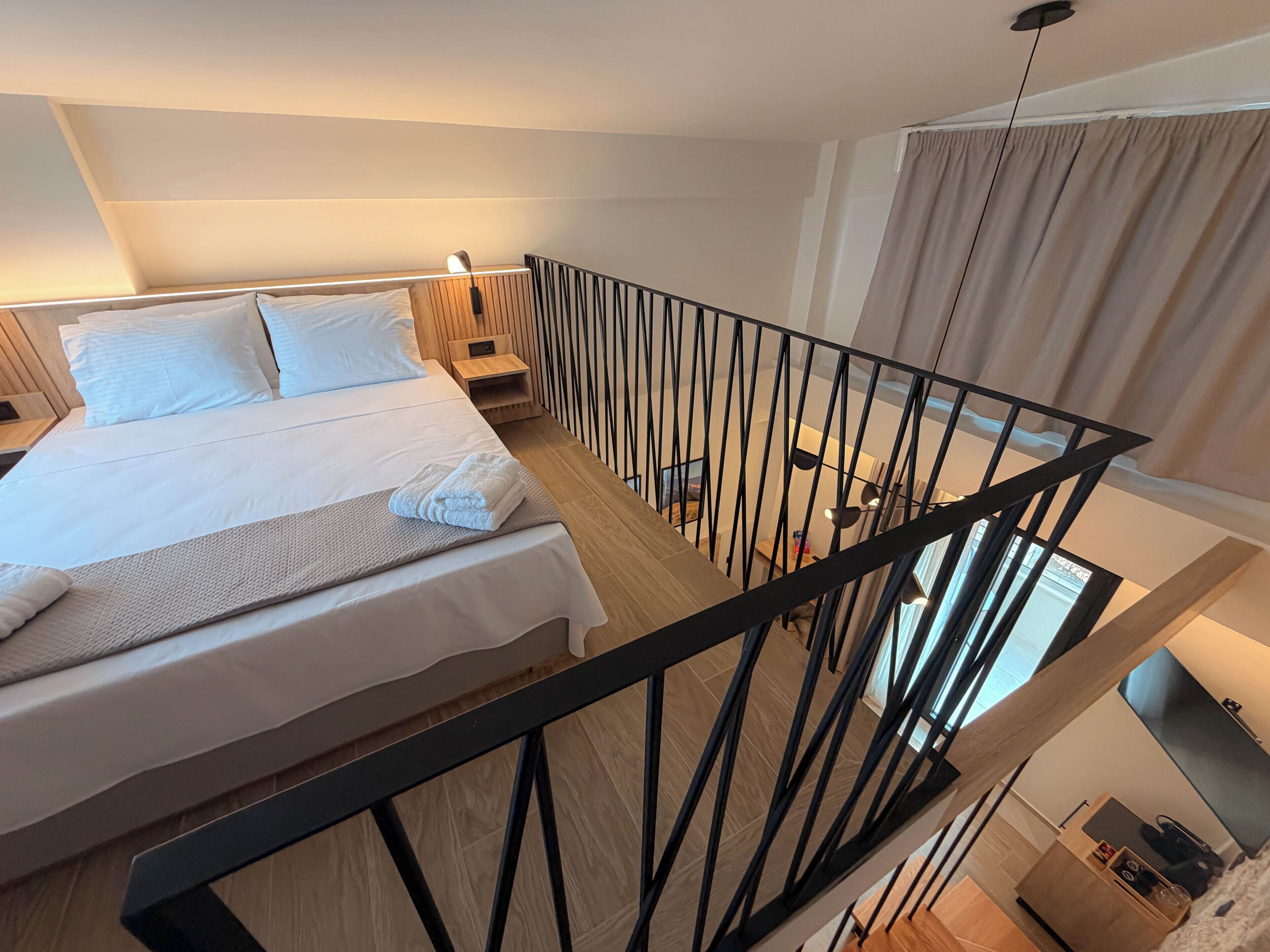 Una habitación de hotel moderna con una cama grande, cabecero de madera, una mesita de noche, barandilla de metal negro y vistas de la ciudad a través de la ventana.