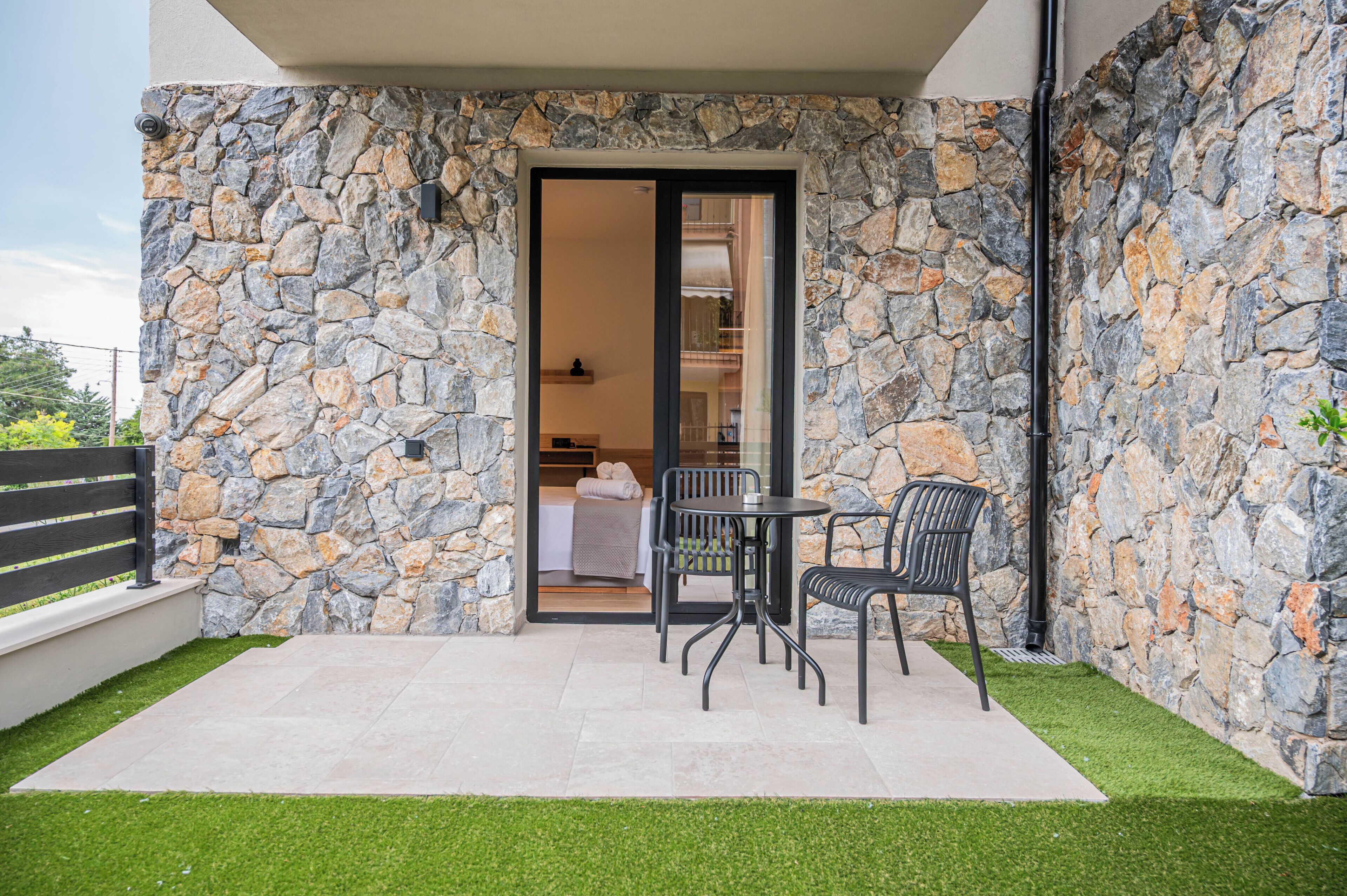 Pared exterior de piedra, una puerta de vidrio que da a una habitación con cama y mesa, y un pequeño patio con sillas y césped artificial.